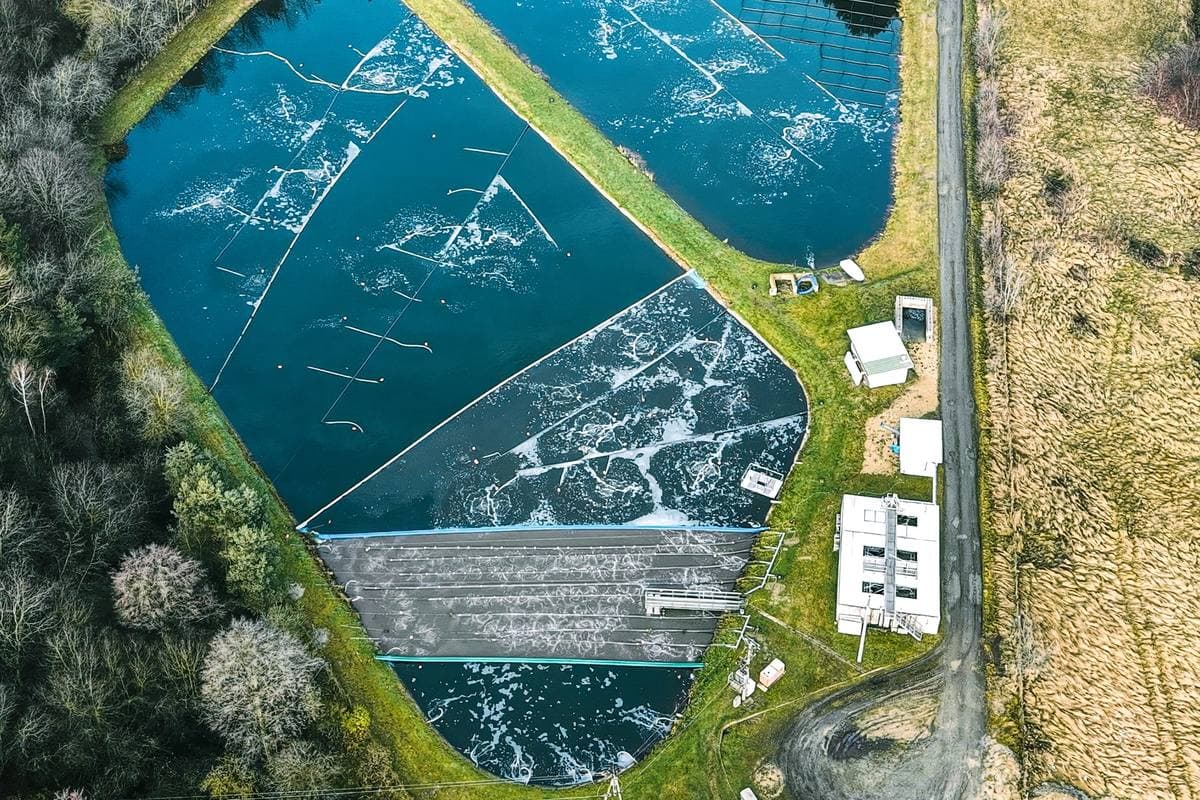 Aerial view of a multi-cell aerated wastewater lagoon, its surface broken by aeration, with grass berms dividing the cells and a small control building alongside.